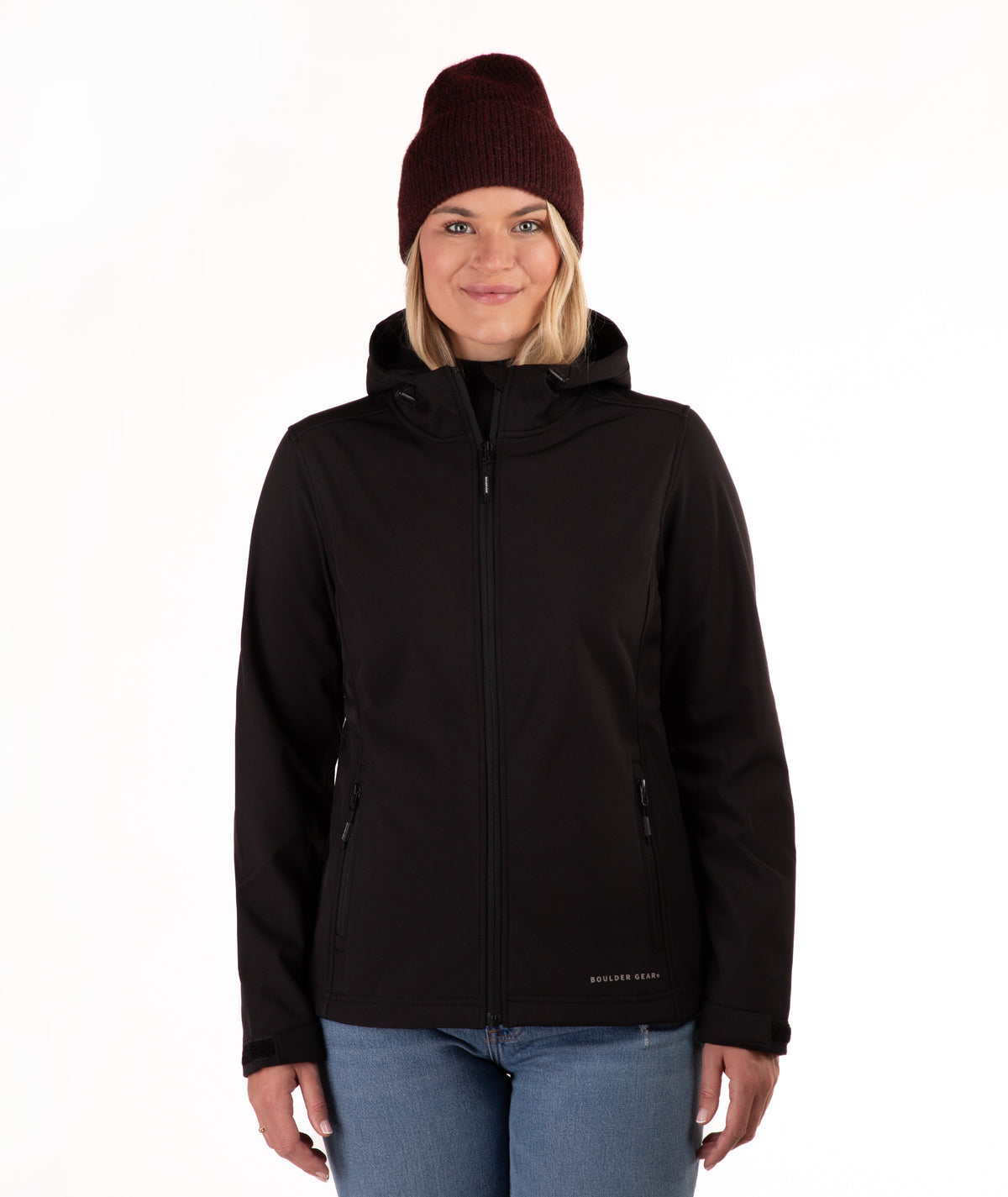 A woman wearing a black hooded zip-up jacket, blue jeans, and a dark burgundy knit beanie, standing against a plain white background.