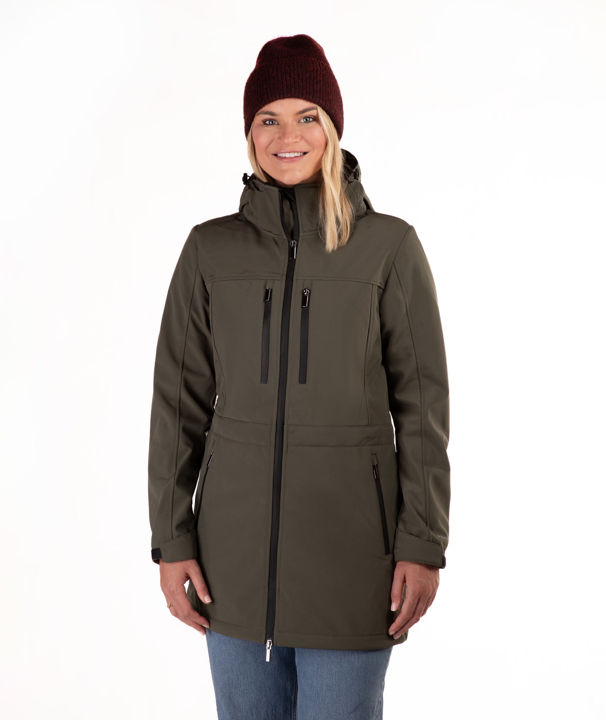 A woman wearing a dark olive green hooded softshell jacket with black zippers, blue jeans, and a burgundy knit beanie, standing against a plain white background.