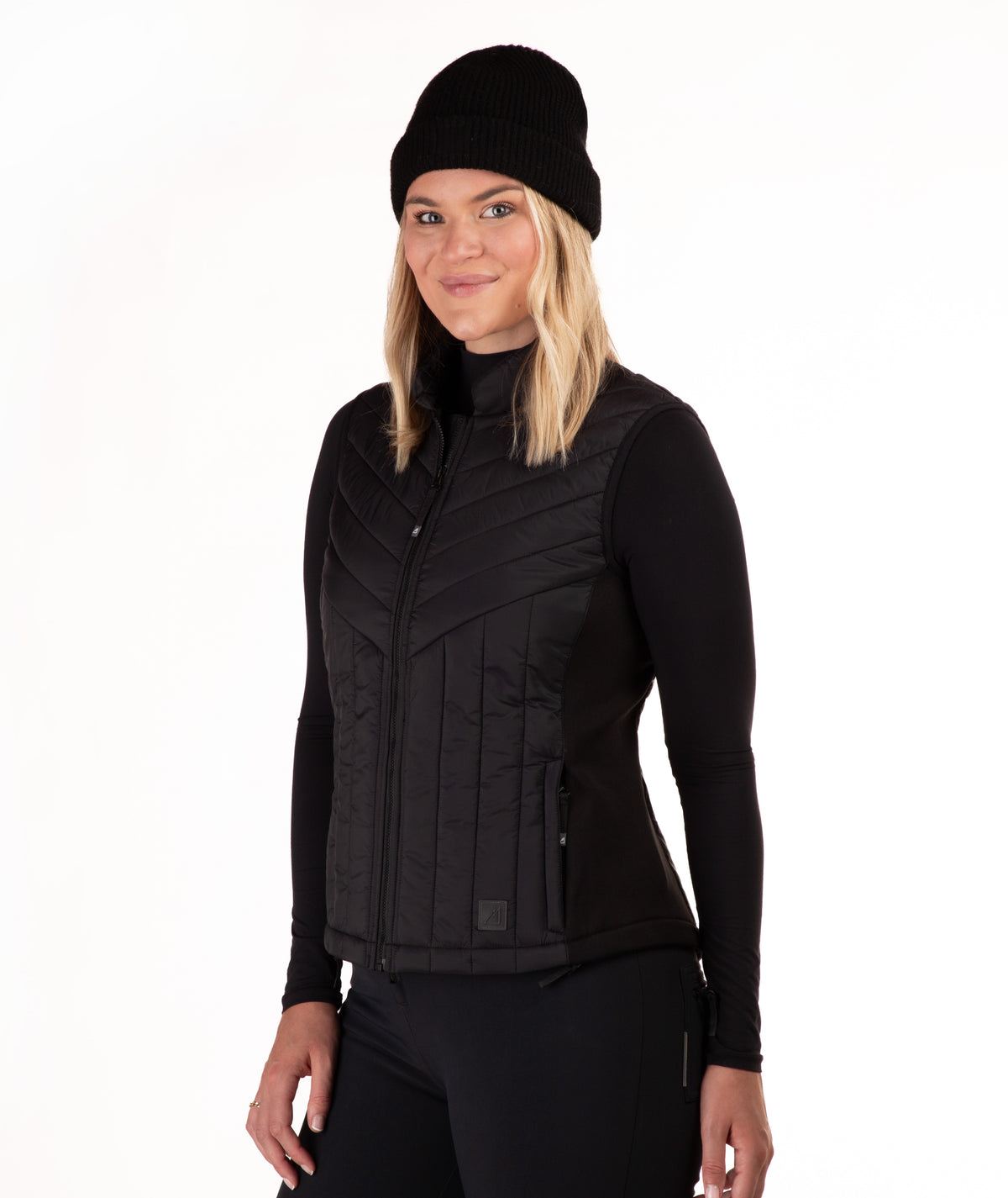A woman wearing a black zip-front quilted vest over a black long-sleeve top and black beanie, photographed against a plain white studio background.