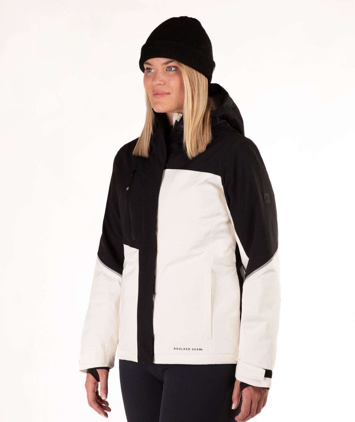 A woman wearing a black and off-white color-blocked ski jacket, a black beanie, and black pants, photographed against a clean white studio background.