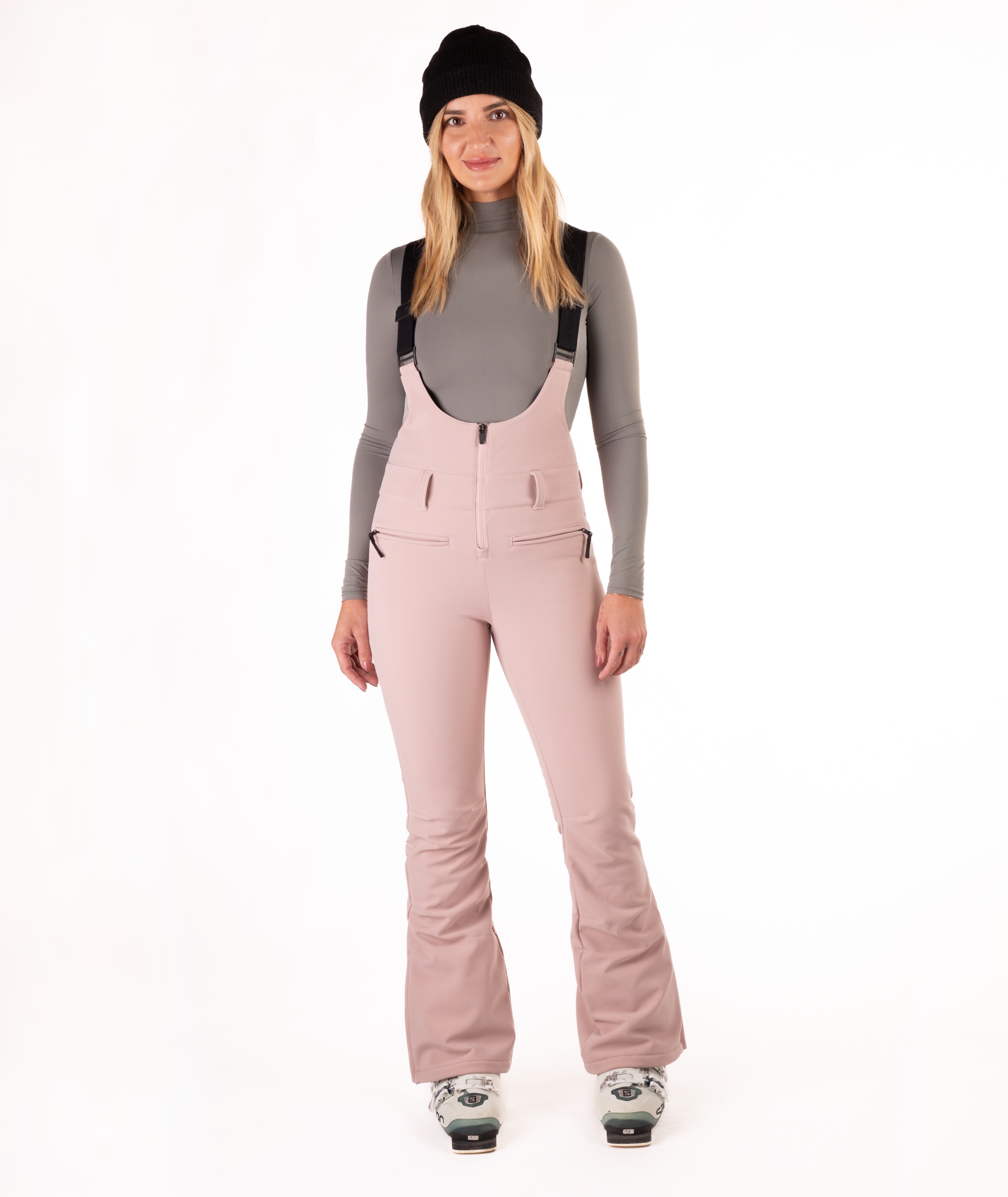 Woman wearing pink ski bib overalls with a gray top on a white background
