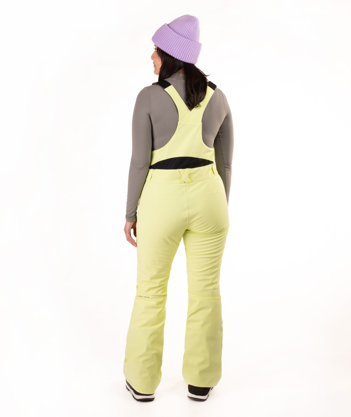 A model shown from the back wearing light yellow-green ski bibs, a gray long-sleeve top, a purple knit beanie, and black and white boots, set against a plain white studio background.