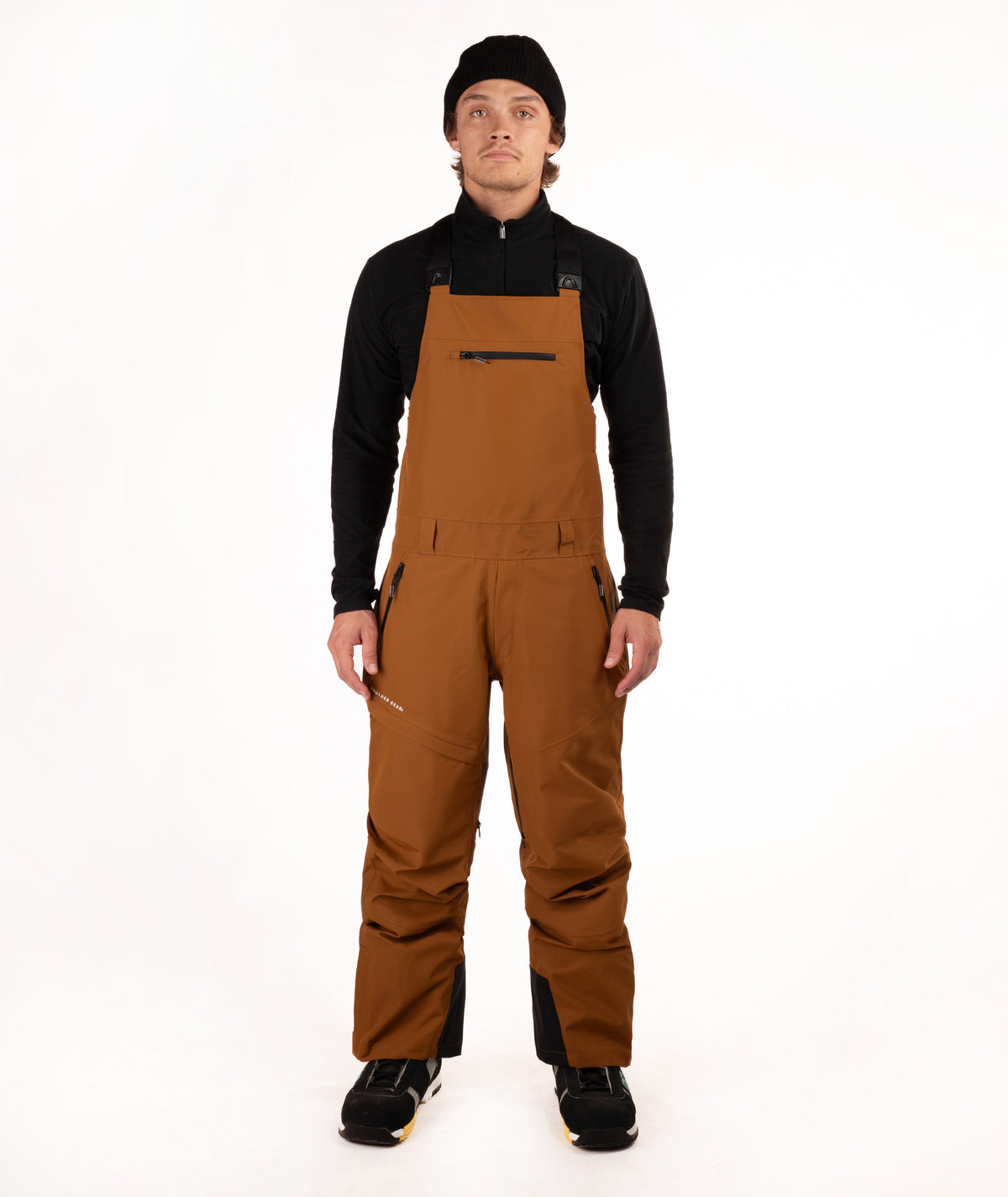 A male model wearing brown snow bib overalls with black panels, a black long-sleeve shirt, and a black beanie, standing against a solid white studio background.