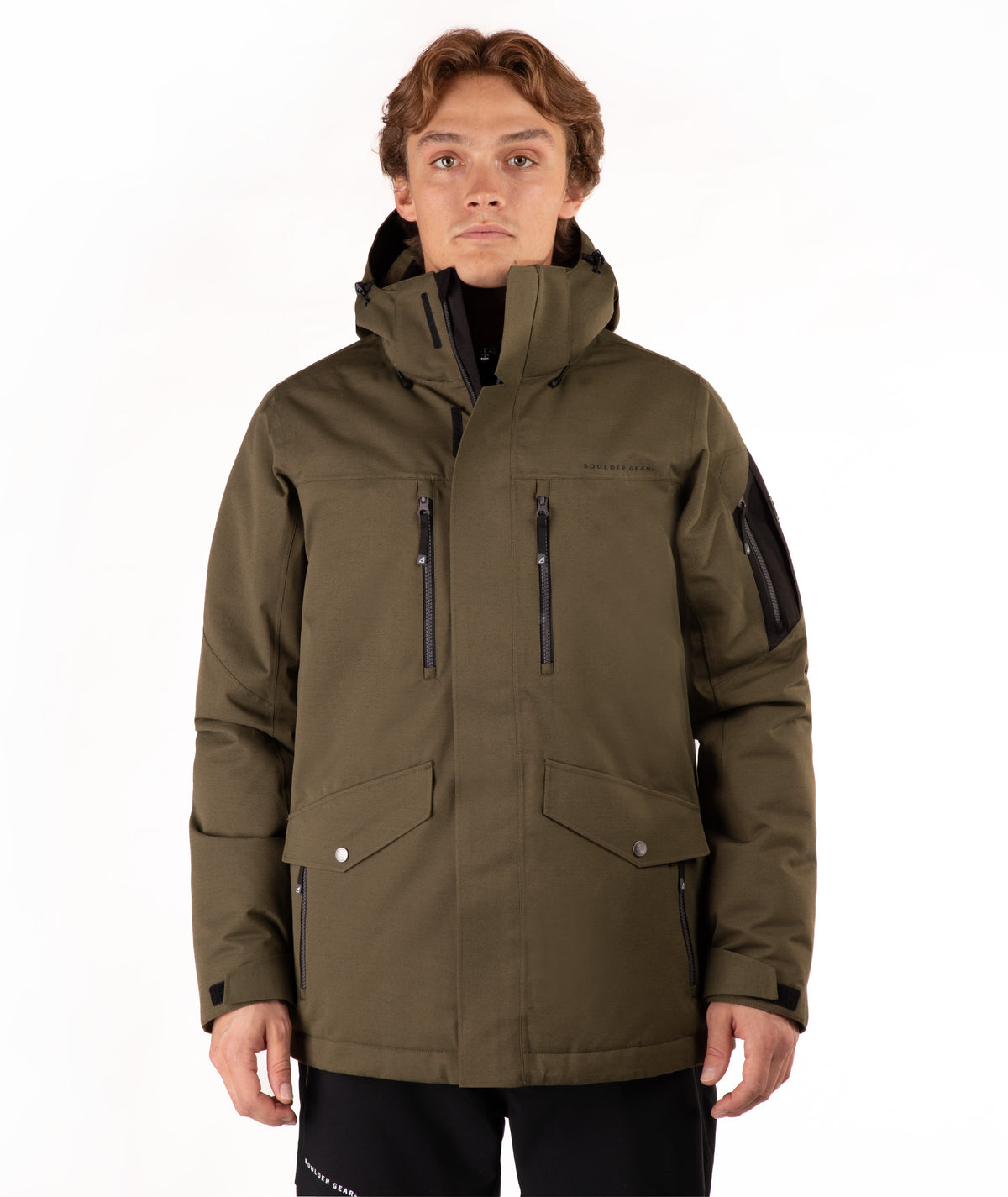 A man wearing an olive green hooded outdoor jacket with black zippers and black pants, photographed against a plain white studio background.