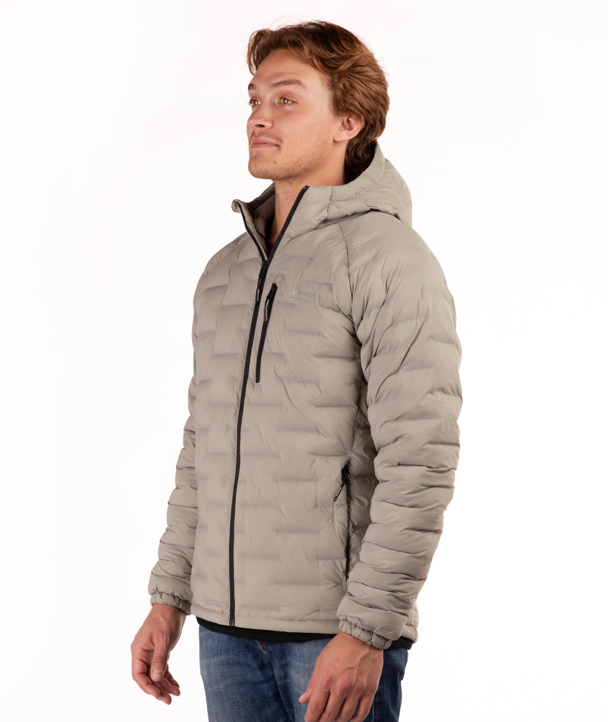 A man wearing a light taupe hooded puffer jacket with black zippers, shown in a side profile against a plain white background.