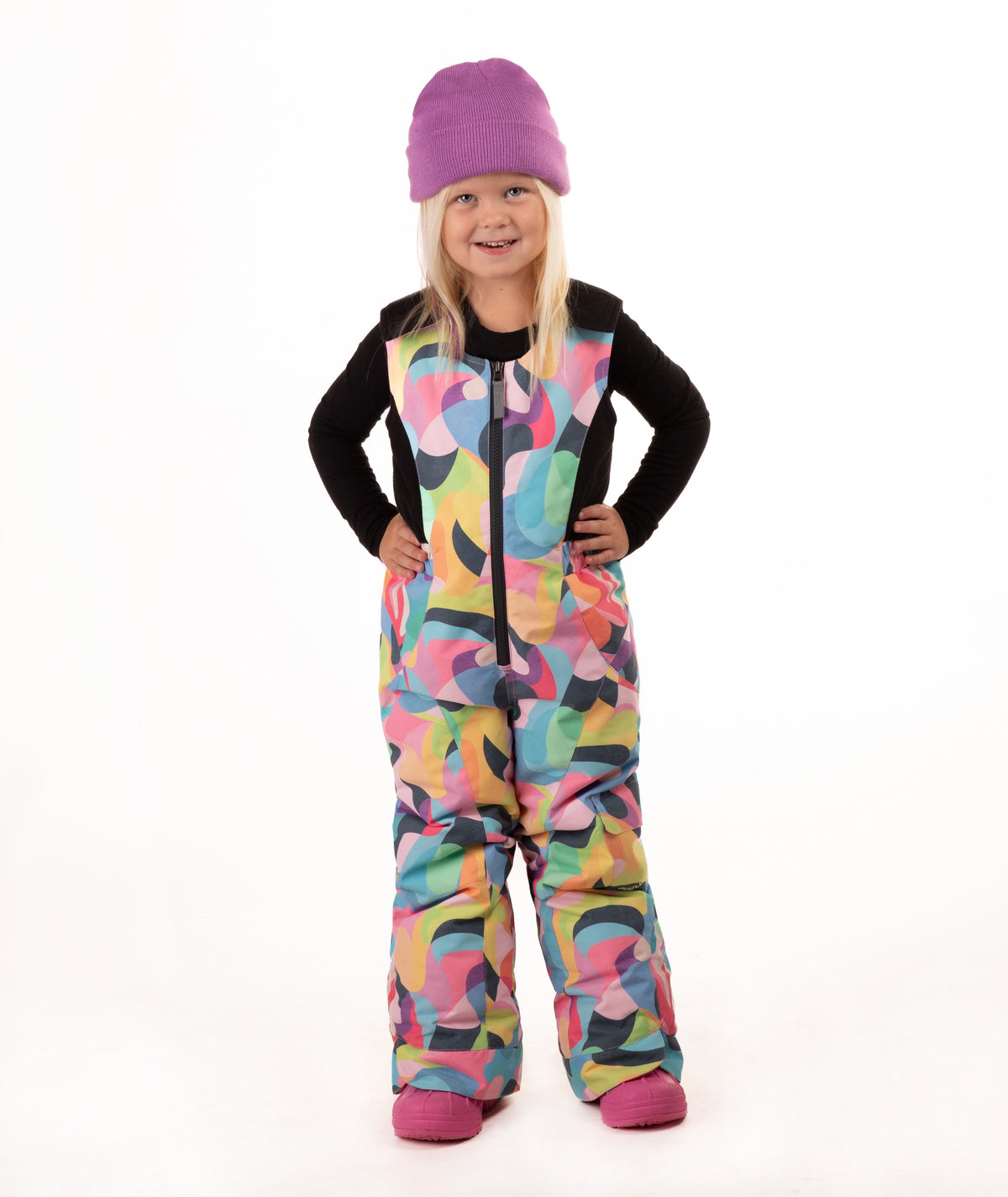 A young child wearing a black long-sleeve top and a colorful abstract print snowsuit with a purple beanie and pink boots, standing against a white studio background.