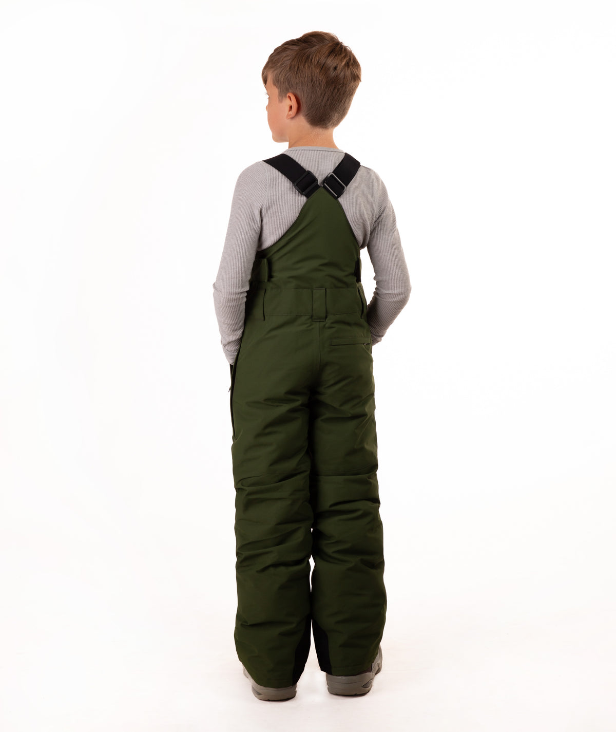 A child viewed from the back wearing dark olive green insulated bib overalls over a light gray long-sleeve shirt, standing against a plain white studio background.