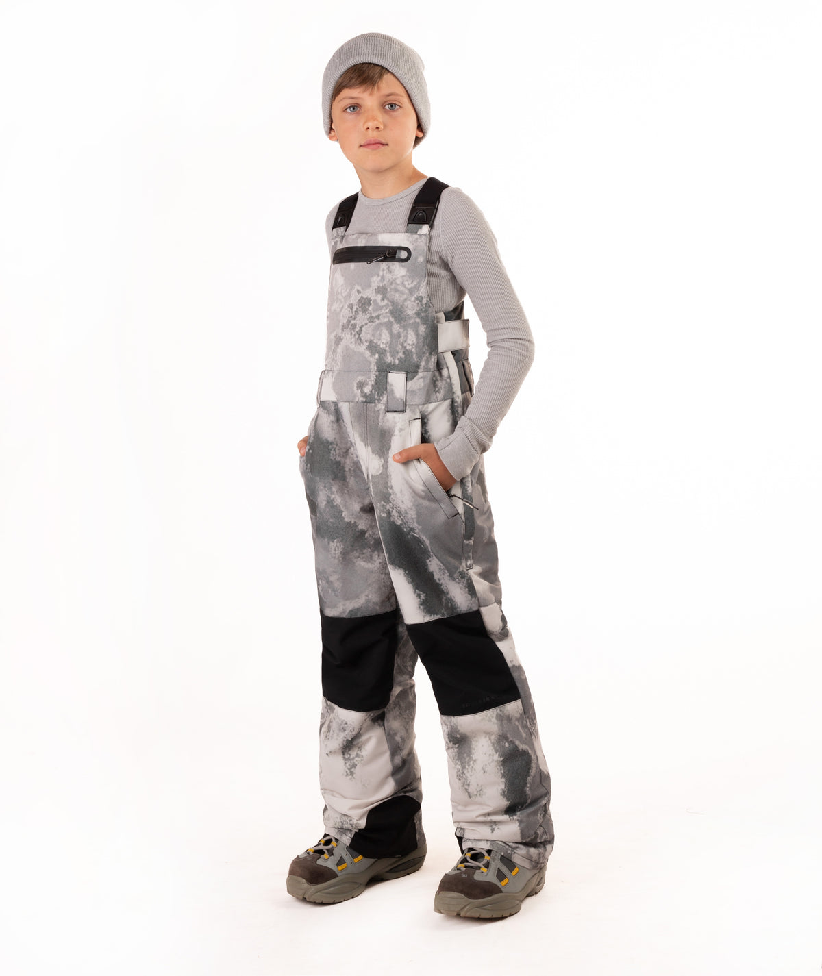 A child wearing gray and black camouflage snow bib overalls with black knee patches, a gray long-sleeve shirt, a gray beanie, and winter boots, standing against a white studio background.