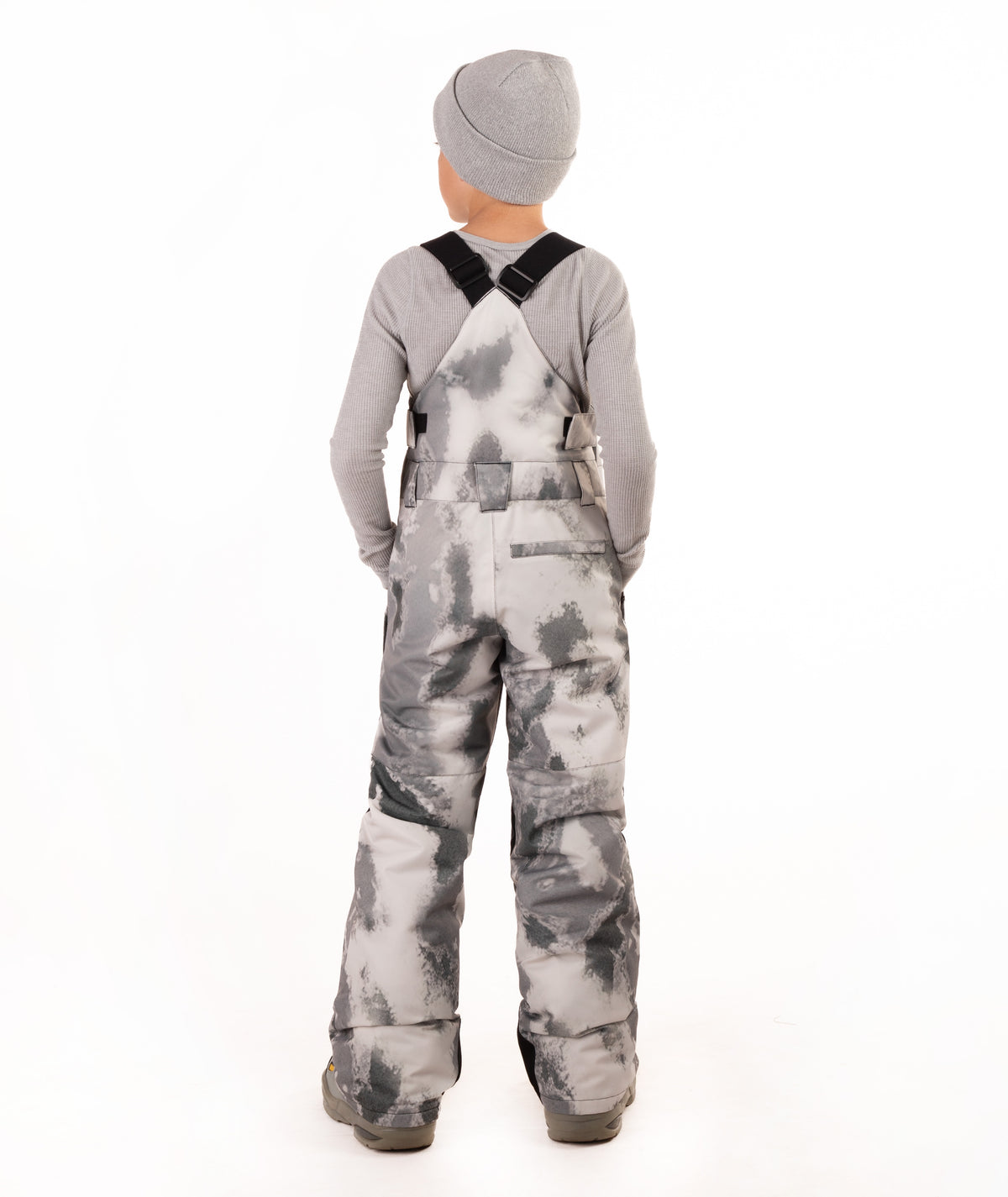 A child viewed from the back wearing gray and white tie-dye snow bib overalls, a gray long-sleeve shirt, and a gray beanie on a white studio background.