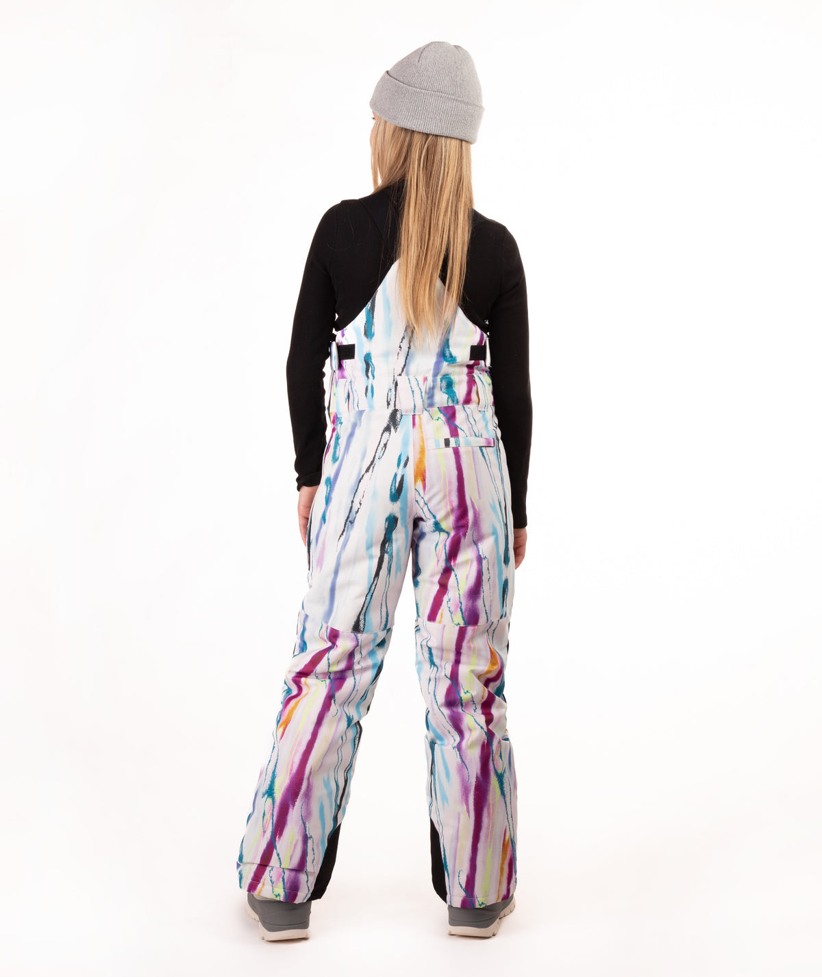 A model viewed from behind wearing a gray beanie, a black long-sleeve shirt, and white snow bibs with a colorful paint-splatter pattern in purple, blue, orange, and yellow, set against a plain white studio background.
