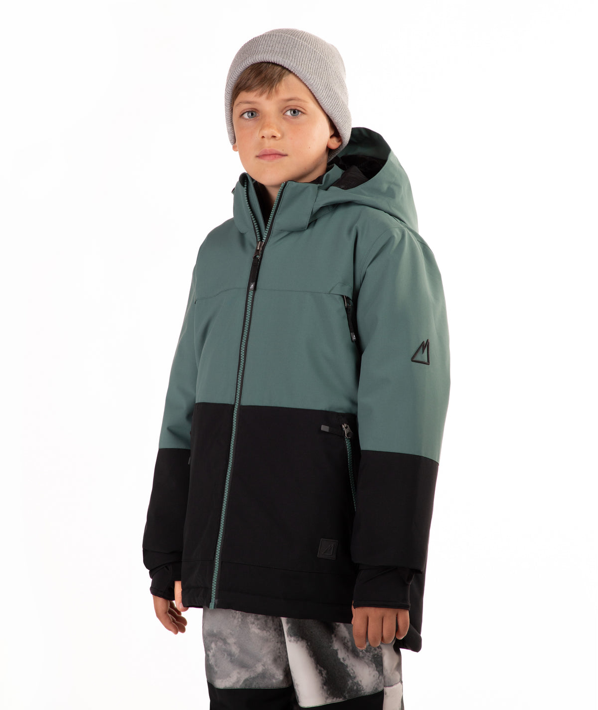 A child wearing a color-blocked ski jacket in sage green and black, paired with a gray beanie and patterned ski pants, standing against a plain white background.