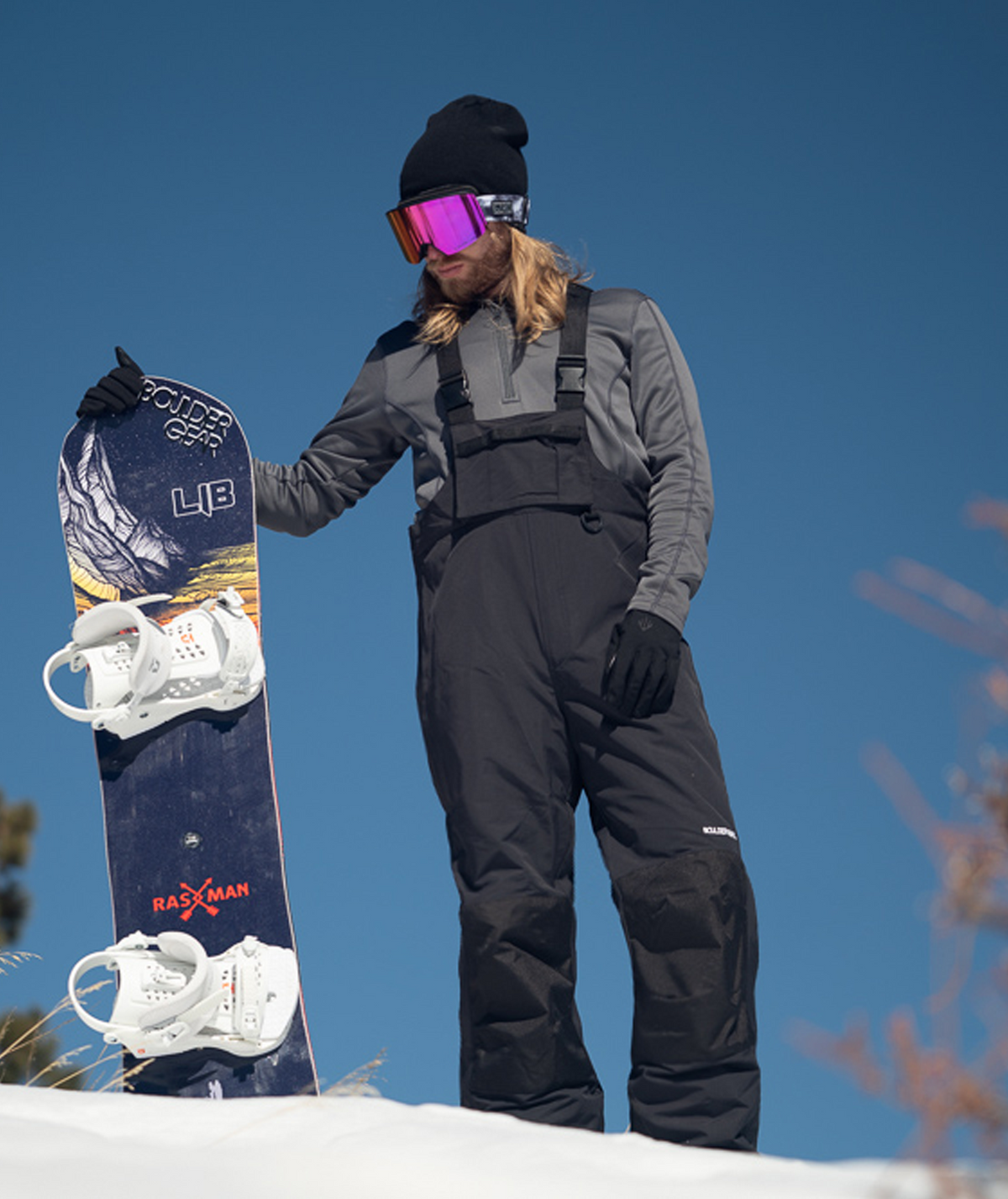 man on a snowy mountain wearing black insulated bibs with front cargo pocket.