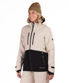 A woman wearing a beige and black snowboard jacket, a tan beanie, and beige snow pants, standing against a solid white background.