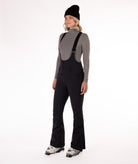 Woman wearing black ski bib overalls with a gray top on a white background