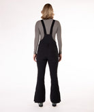 back of a Woman wearing black ski bib overalls with a gray top on a white background