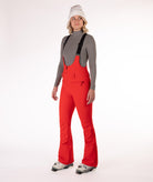 Woman wearing red ski bib overalls with a gray top on a white background