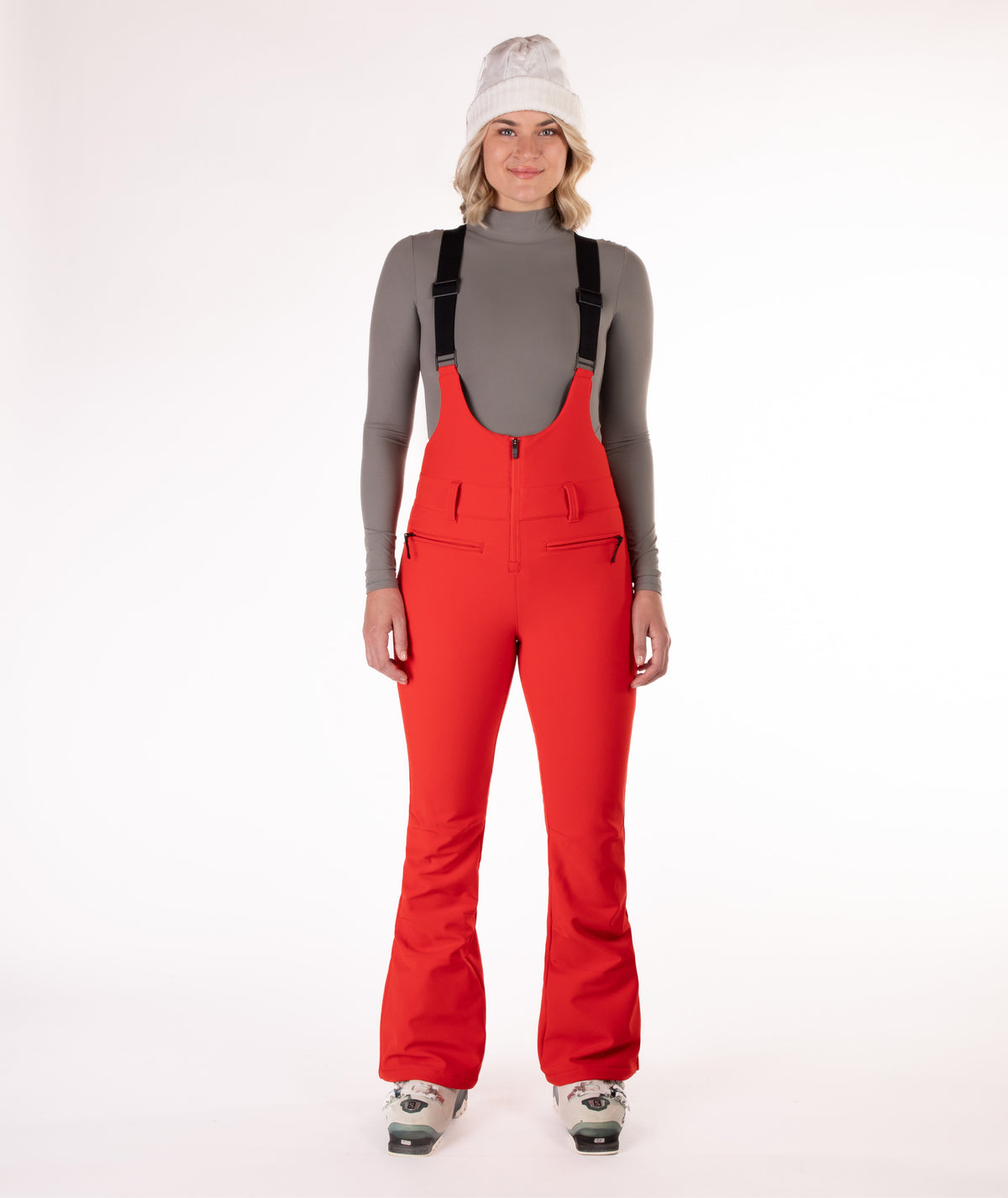 Woman wearing red ski bib overalls with a gray top on a white background