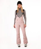 Woman wearing pink ski bib overalls with a gray top on a white background