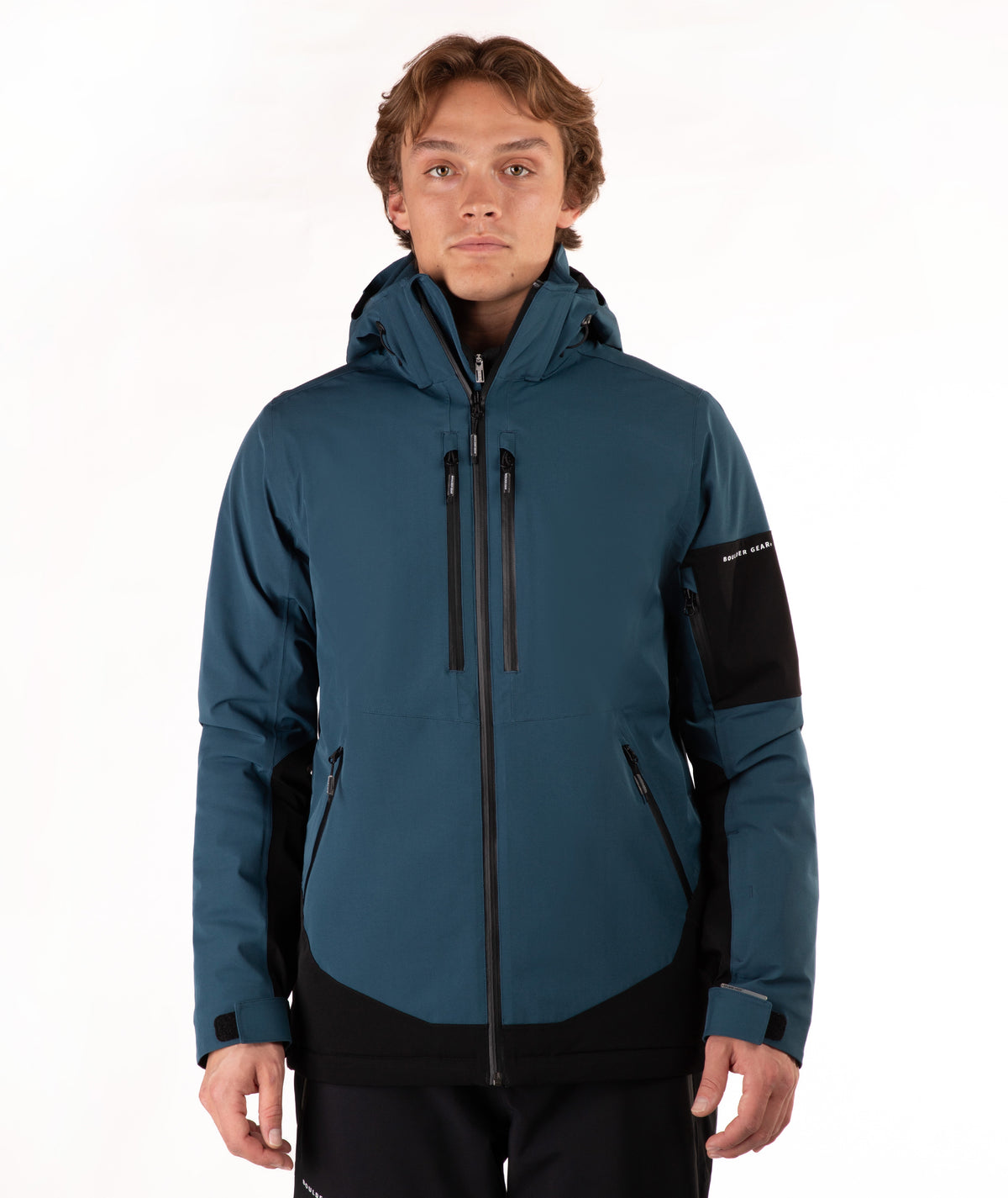 A man wearing a dark teal and black hooded technical jacket with a full front zipper, zippered hand pockets, and a black sleeve patch, photographed against a plain white studio background.