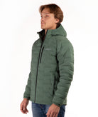 A man wearing a green hooded puffer jacket with a black front zipper and a small white logo on the left chest, paired with blue jeans, against a plain white background.