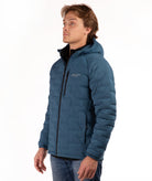 A man wearing a teal blue hooded puffer jacket paired with blue jeans, against a plain white background.