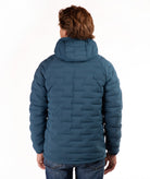 A person viewed from the back wearing a quilted hooded puffer jacket in a blue-teal color, paired with blue jeans, against a plain white studio background.