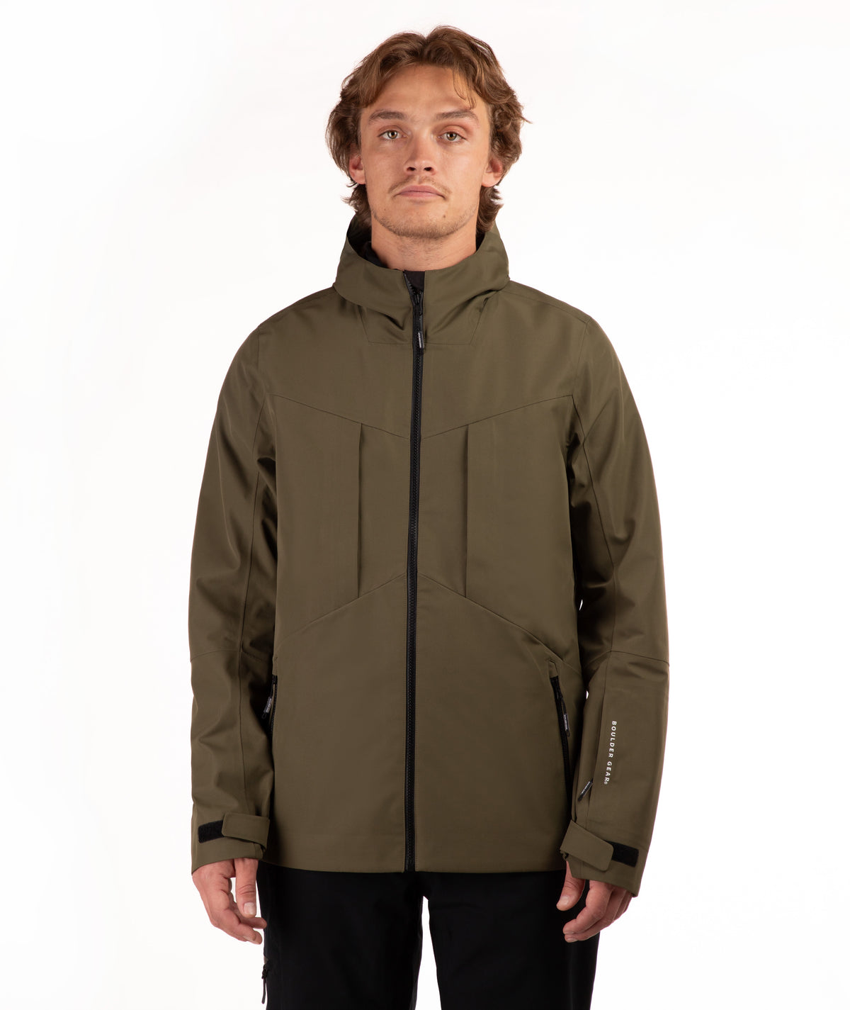 A man wearing an olive green technical outdoor jacket with black zippers and black pants, standing against a plain white studio background.