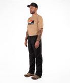 Man wearing a tan Boulder Gear t-shirt and black rain pants on a white background