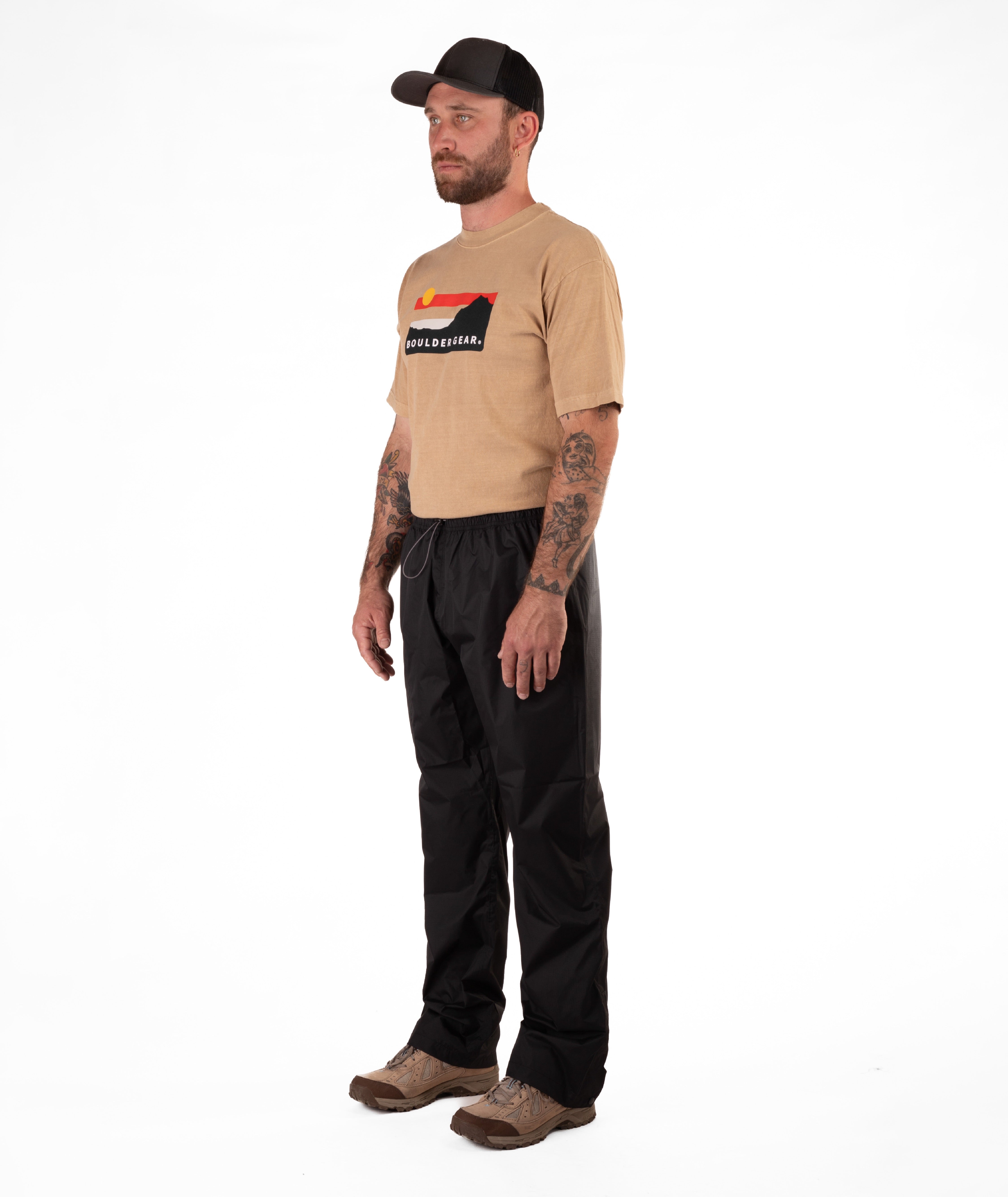 Man wearing a tan Boulder Gear t-shirt and black rain pants on a white background