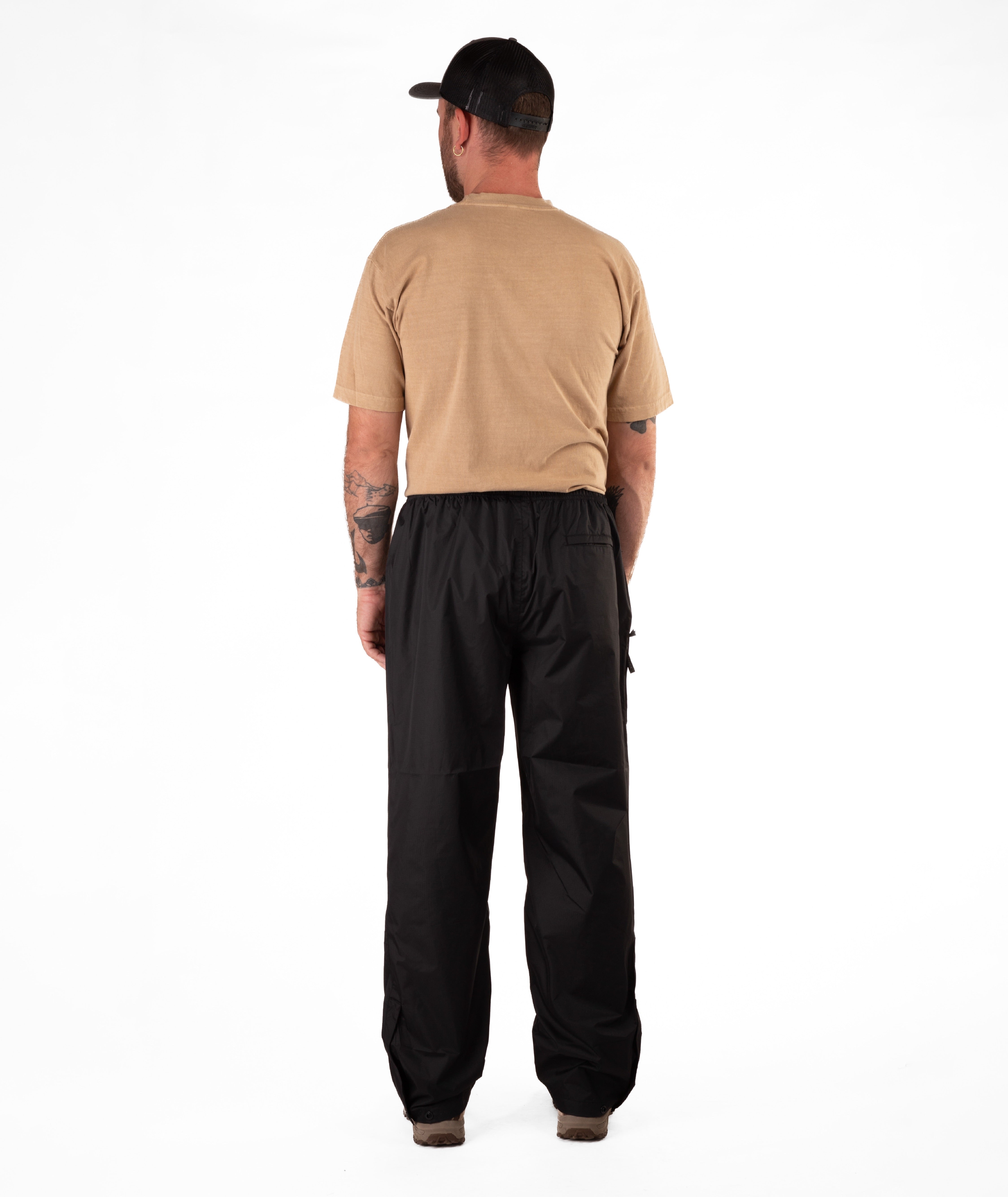 back of a Man wearing a tan Boulder Gear t-shirt and black rain pants on a white background