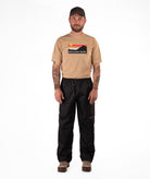 Man wearing a tan Boulder Gear t-shirt and black rain pants on a white background