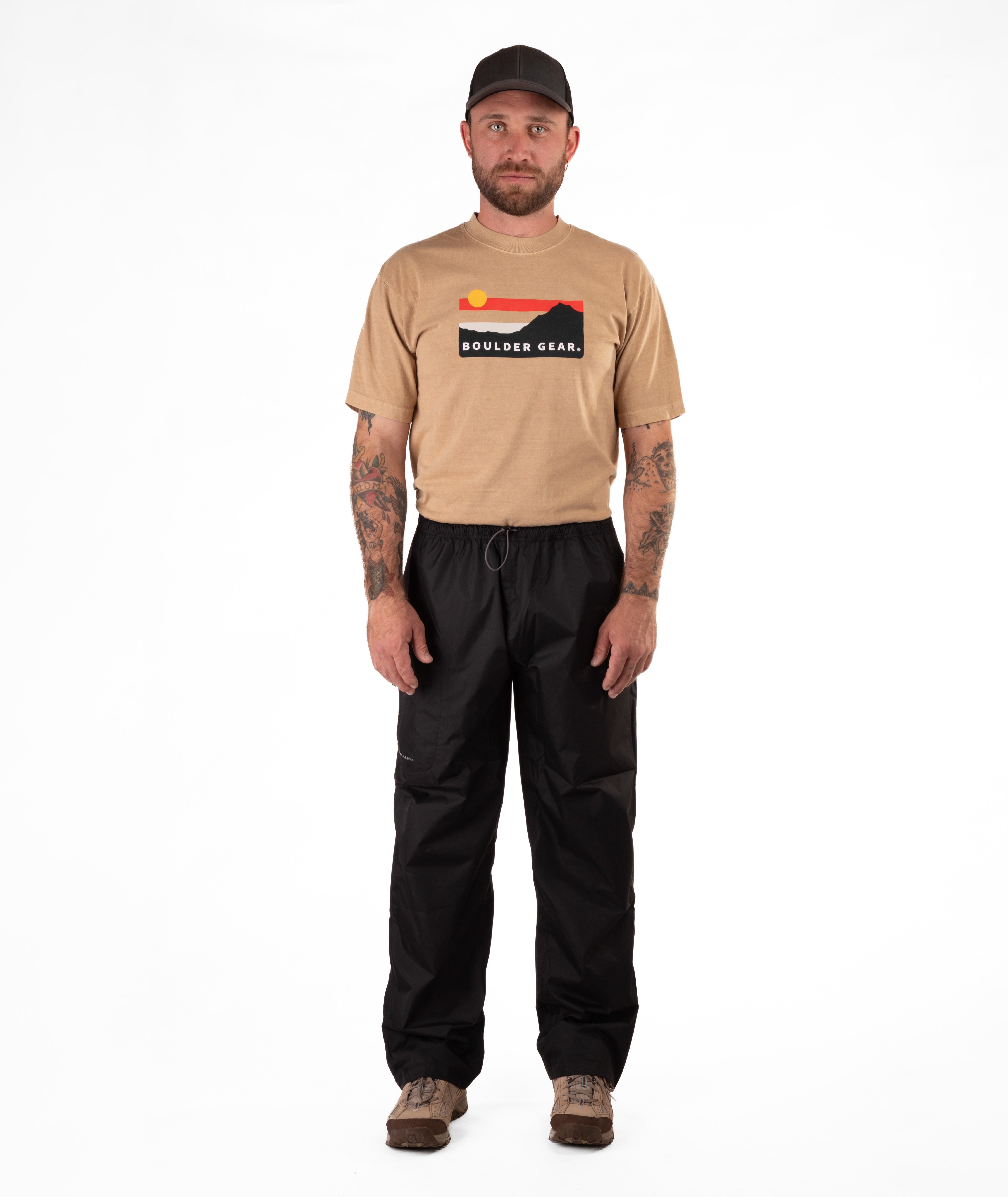 Man wearing a tan Boulder Gear t-shirt and black rain pants on a white background