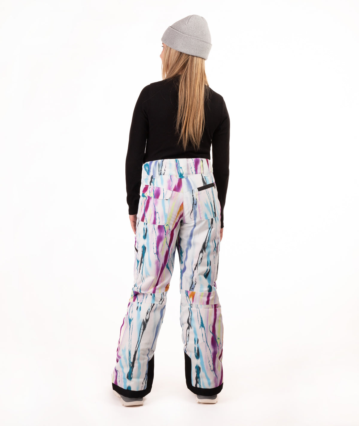 A model shown from the back wearing  snow pants with a colorful paint-splatter pattern, a black long-sleeve top, and a gray beanie against a white studio background.