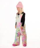 A young girl wearing a pastel rainbow tie-dye snow bib over a black long-sleeve shirt, a pink beanie, and pink boots, standing against a white studio background.