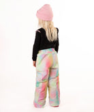 A child shown from the back wearing a pink beanie, a black long-sleeve top, and snow bib with a pastel tie-dye pattern in pink, green, blue, and yellow, against a plain white background.