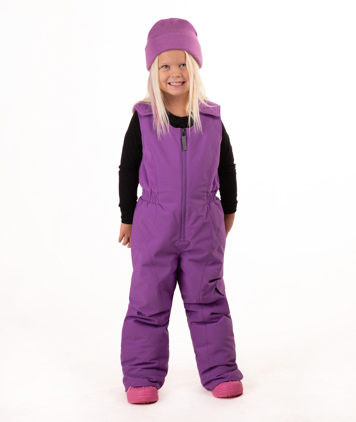 A young child wearing a purple snow bib over a black long-sleeve shirt, a matching purple beanie, and pink boots, standing against a plain white studio background.