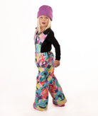 A young child wearing a black long-sleeve top and a colorful abstract print snowsuit with a purple beanie and pink boots, standing against a white studio background.