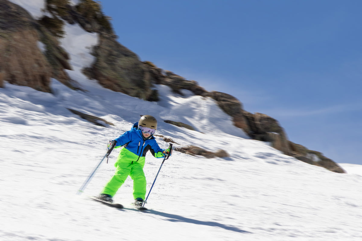 A child skier in a bright blue and neon green ski outfit glides down a snowy mountain slope under a clear blue sky, with rocky cliffs in the background.