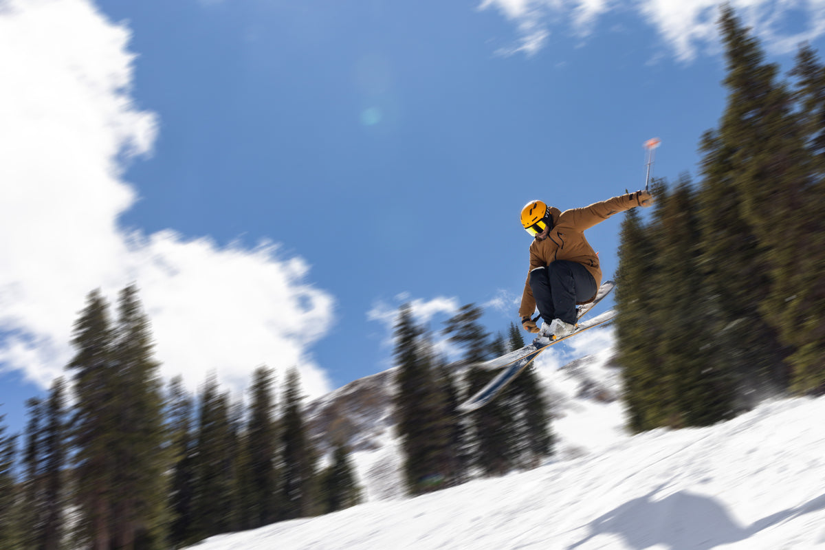 A skier mid-air performing a jump on a snowy mountain slope, surrounded by tall evergreen trees and a bright blue sky with white clouds.