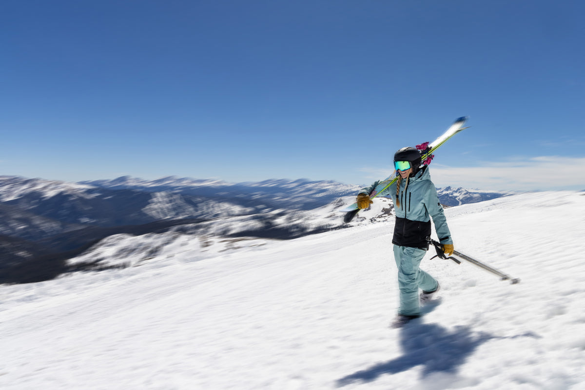 A skier in a light green and black ski outfit stands on a snowy mountain slope under a clear blue sky, holding ski poles and carrying skis over their shoulder. Snow-covered mountains are visible in the background.