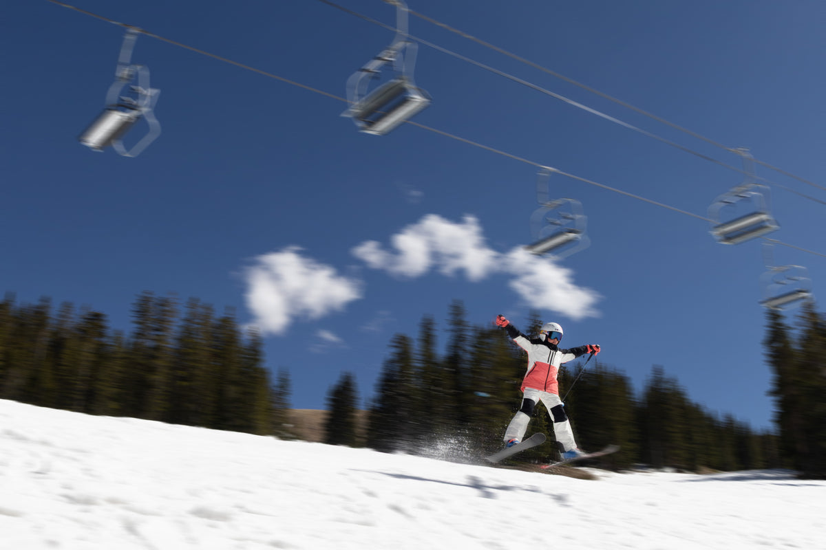 A skier in a white, coral, and black outfit jumps on a snowy slope under a blue sky with clouds and ski lifts overhead.